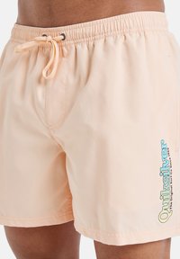 Bañadores de color melocotón con cintura elástica y cordón. Presentan un logotipo azul en la parte inferior izquierda. Textura de tela suave.