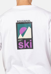 Bílý tričko s grafickým potiskem hor a fialového kruhu. Text zahrnuje "ROSSIGNOL" a "ski" ve výrazných fialových písmenech.