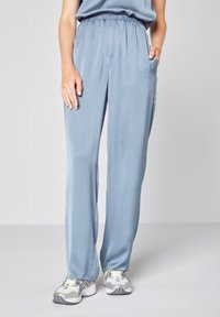 Světle modré, lehké kalhoty s elastickým pasem, dvěma bočními kapsami a hladkým povrchem, spárované s šedými a žlutými sportovními botami.