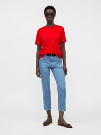 Personne portant une chemise rouge vif à manches courtes, un jean bleu clair avec une ceinture noire, des ballerines à imprimé léopard et des lunettes de soleil teintées marron, debout.