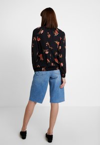 Donna in piedi di spalle, con indosso una blusa nera a fiori con maniche lunghe, pantaloni corti in denim blu fino al ginocchio e scarpe nere su uno sfondo bianco.