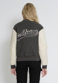 Grå uld bomberjakke med cremede quiltede ærmer, som har et stort broderet "Mustang" logo på ryggen. Ribbet kant og manchetter.