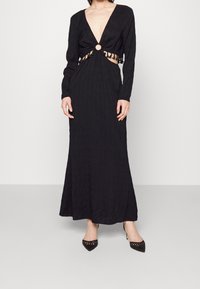 Robe noire à manches longues avec un profond décolleté en V, détail découpé à la taille et ornements circulaires. Tissu texturé, corsage ajusté et jupe fluide.