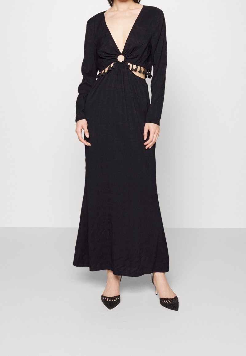 Robe noire à manches longues avec un profond décolleté en V, détail découpé à la taille et ornements circulaires. Tissu texturé, corsage ajusté et jupe fluide.