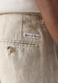 Bežist linased püksid küljekotiga, väike valge silt, millel on "Marc O'Polo" ja ümmargune nööbikaunistus kotil.