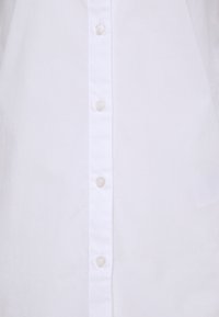 Camicia bianca con bottoni, realizzata in tessuto morbido, con una fila di bottoni trasparenti al centro. Design semplice, senza motivi o dettagli.
