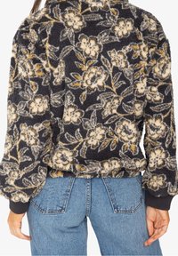 Chaqueta de forro polar en azul marino oscuro con un patrón floral en crema y dorado. Presenta una superficie texturizada y puños acanalados, con un ajuste suelto y corto.