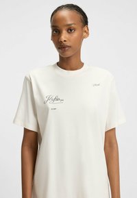 Cremefarvet bomulds T-shirt med korte ærmer og subtil trykt tekst i sort. Løs pasform med en glat tekstur og minimalistisk design.