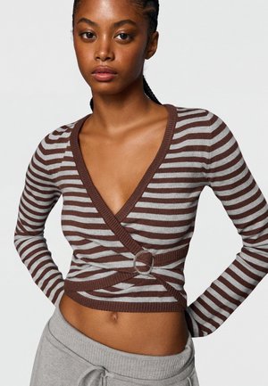 Jeune femme portant un pull cache-cœur rayé marron et gris avec boucle et un pantalon gris à cordon sur un fond uni.