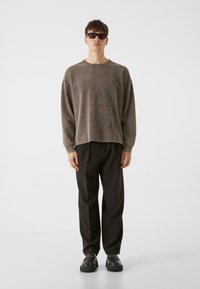 Suéter de gran tamaño y textura color marrón claro con un cuello ancho, combinado con pantalones negros holgados y zapatos negros gruesos. El modelo se mantiene erguido.