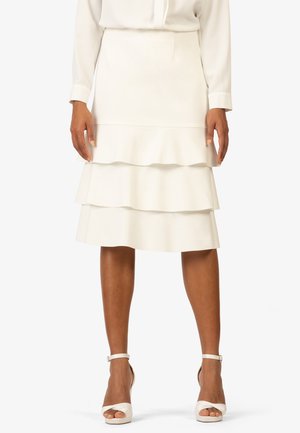 Jupe blanche à volants avec trois couches froncées, fabriquée à partir d'un tissu lisse. Associée à des sandales à talons blancs avec des sangles de cheville.