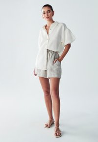 Linnen short-sleeve shirt in crème, met een knoopsluiting aan de voorkant en een relaxed fit. In combinatie met lichtgrijze shorts met koord. Schoenen zijn strappy sandalen.