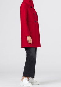 Cappotto rosso in misto lana con colletto alto, design doppiopetto, bottoni neri e tasche laterali. Abbinato a jeans scuri e sneakers bianche.