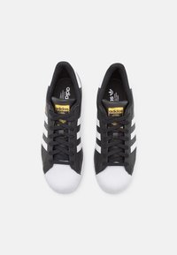 Een paar zwarte Adidas sneakers met witte shell-toes en strepen, zwarte veters en gouden logopatches op de tong, gezien van bovenaf.