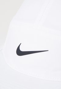 Hvit caps laget av glatt stoff, med en buet skjerm. En svart Nike-logo er tydelig vist foran. Enkle sømdetaljer synlige.