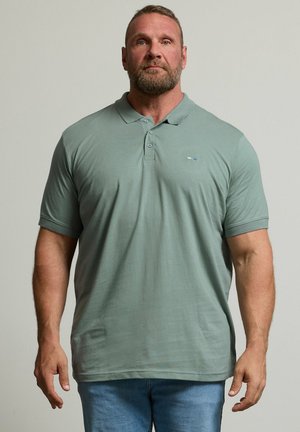 Muskelbepackter Mann mit kurzen Haaren und Bart, der ein hellgrünes Poloshirt und blaue Jeans trägt, steht vor einem schlichten grauen Hintergrund.