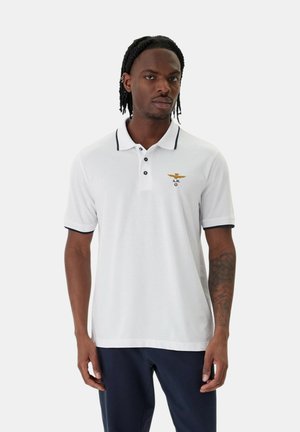 Uomo che indossa una polo bianca con bordature nere e stemma dorato ricamato sul petto, abbinata a pantaloni blu navy, in piedi davanti a uno sfondo semplice.