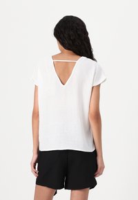 Biały lniany top z krótkim rękawem, z wycięciem na plecach w kształcie litery V i subtelną fakturą. Połączony z czarnymi szortami, podkreślającymi casualowy styl.
