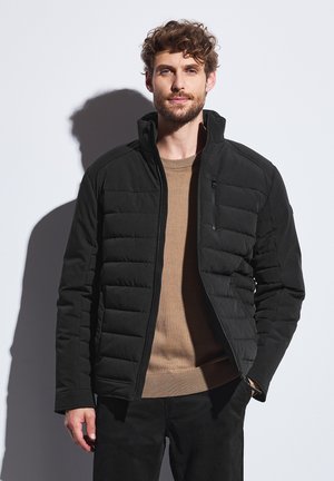Chaqueta negra aislante con diseño acolchado y cuello alto. Presenta un bolsillo con cremallera en el pecho y textura suave. Lleva puesta sobre un suéter color beige.