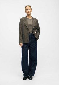 Blazer brown oversize sopra una camicia grigia, abbinato a jeans in denim scuro a gamba larga e scarpe nere. Il blazer presenta revers affilati e un solo bottone.