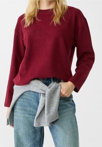 Vrouw in een lang mouwen bordeauxrood sweater, blauwe jeans, en een grijze sweater om haar taille geknoopt, staand tegen een effen achtergrond.