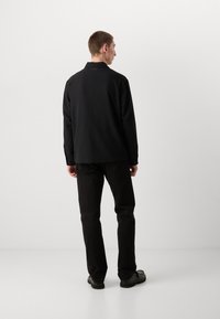 Black long-sleeve shirt with a textured surface and classic collar, paired with black pants and sturdy black shoes, viewed from behind.