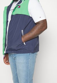 Marineblauer und grüner leichter Weste mit Reißverschluss, seitlichen Taschen und Tommy Jeans-Logo auf der Brust. Über einem weißen T-Shirt getragen.