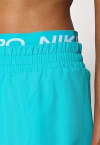 Shorts de sport turquoise avec une taille texturée présentant le logo Nike en blanc. Tissu lisse, taille élastique et détails froncés.