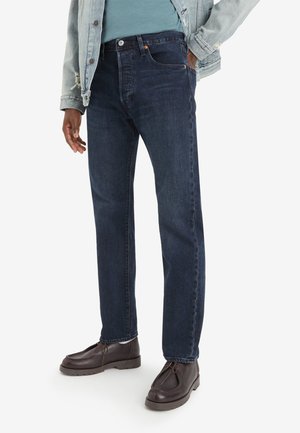 Jeans in denim blu scuro con una vestibilità dritta, caratterizzati da un classico design a cinque tasche e chiusura a bottoni. Indossati con una giacca in denim chiaro e scarpe marroni.