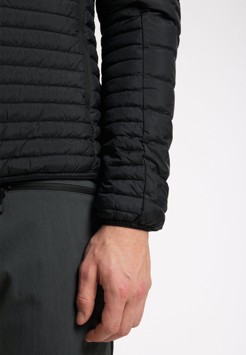 Chaqueta negra aislante con diseño acolchado, textura suave y puños elásticos, combinada con pantalones gris oscuro. Primer plano del brazo y la mano.