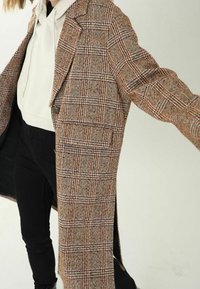 Ruskea ruudullinen pitkä takki, jossa on rakenteellinen muotoilu, notched-läppä, yksittäinen napitus ja sivutaskut. Sen alla on näkyvissä valkoinen huppari.