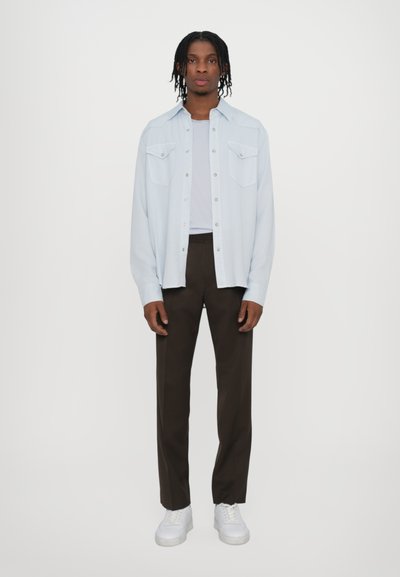 Svetlo modrá košeľa s gombíkmi a dvoma vrecami na hrudi, vrstvená nad šedým tričkom, skombinovaná s hnedými nohavicami a bielymi teniskami.