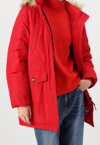 Rød dunjakke med hætte foret med pels, forlommer foran og lynlås lukning, båret over en rød sweater, kombineret med blå jeans.