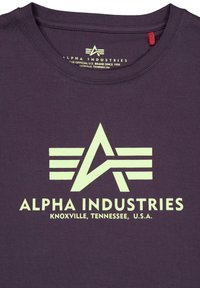 Lila pamut póló nagyméretű, tükröződő logóval, amelyen egy 'A' és szárny design található, alatta a "ALPHA INDUSTRIES" felirat és a helyszín adatai.