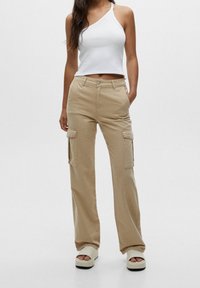 Vrouw in beige cargobroek, wit één-schouder crop top en witte plateau-sandalen, staand met één hand in de zak.