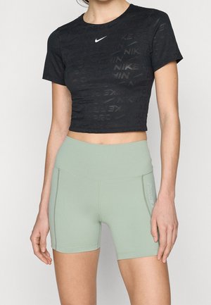Kobieta ubrana w czarny krótki T-shirt Nike oraz jasnozielone, wysokie spodenki rowerowe Nike na jednolitym tle.