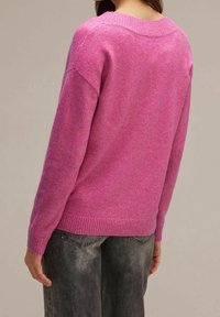 Femme portant un pull en tricot rose à manches longues et un jean noir délavé, vue de dos sur un fond uni.