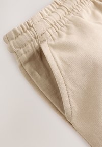 Gros plan d'un pantalon en tissu beige montrant la ceinture élastique et le détail des coutures de la poche latérale.