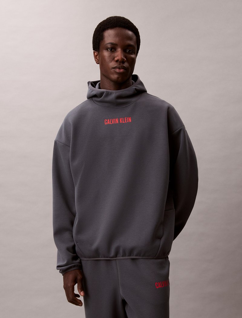 Felpa grigia con collo alto e polsini a costine, caratterizzata da un audace logo rosso "CALVIN KLEIN" sul petto, realizzata in tessuto morbido.