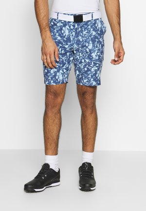Homme portant un short à motifs floraux bleus et blancs avec une ceinture blanche, des chaussettes blanches et des chaussures de sport noires.