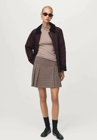 Veste bomber bordeaux avec col noir, haut ajusté beige et jupe plissée à carreaux marron. Des chaussettes noires et des chaussures complètent le look.