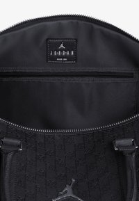 Interior de bolso tote negro con textura satinada suave. Cuenta con un bolsillo con cremallera y una etiqueta de logo en relieve, con un diseño estampado en todo el tejido.