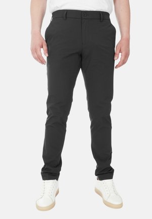 Pantaloni neri realizzati in un materiale liscio e elastico con una vestibilità affusolata, dotati di chiusura con bottone e passanti per cintura. Abbinati a sneaker bianche.