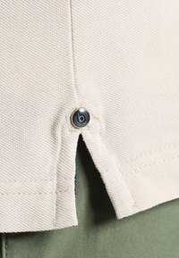 Close-up of a light beige shirt hem with a decorative button showing a logo. The shirt is partially tucked into green fabric below.