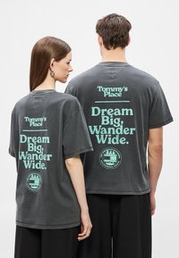 Γκρι υπερμεγέθη κοντομάνικα μπλουζάκια με πράσινα μέντα γράμματα στην πλάτη: "Tommy's Place" και "Dream Big, Wander Wide."