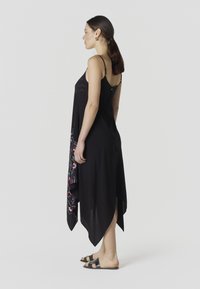 Femme debout de profil portant une robe asymétrique noire à motifs floraux et des sandales noires à enfiler sur un fond uni.