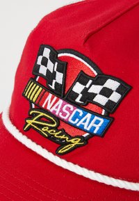 Gorra roja con un diseño bordado que incluye banderas a cuadros, un logotipo circular y el texto "NASCAR Racing" en varios colores. Detalle de ribete blanco.