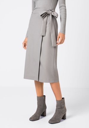 Femme portant une jupe portefeuille gris clair avec un nœud attaché sur le côté, associée à des bottines gris daim et un haut à manches longues côtelé.