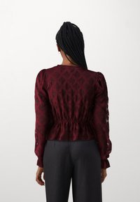 Burgundia blouse tekstuuriga mustriga, pikkade puffvarrukatega ja kogutud vöökohaga. Kaasas tumehallid püksid, vaadatud tagant.