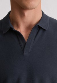 Mężczyzna noszący granatową, fakturowaną koszulę polo z otwartym kołnierzykiem, odsłaniającą część szyi i dolnej części twarzy.